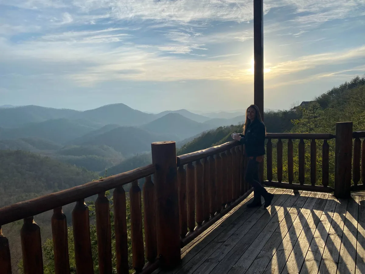 A close-up, inviting scene showing a person holding a cup on a wooden balcony, gazing at the sunrise over expansive mountain landscapes. The composition captures the peaceful solitude and connection with nature possible right at the doorstep, bridging the excitement of outdoor adventure with moments of tranquil reflection.