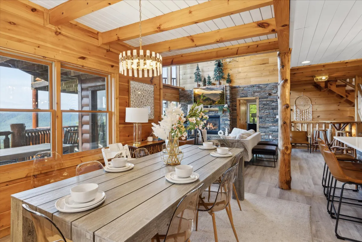 A warm, inviting dining area featuring a large wooden table set for a meal, a glowing chandelier, a stone-surround fireplace, decorative plants, and expansive windows framing the mountain scenery. The spacious layout fosters a sense of togetherness.