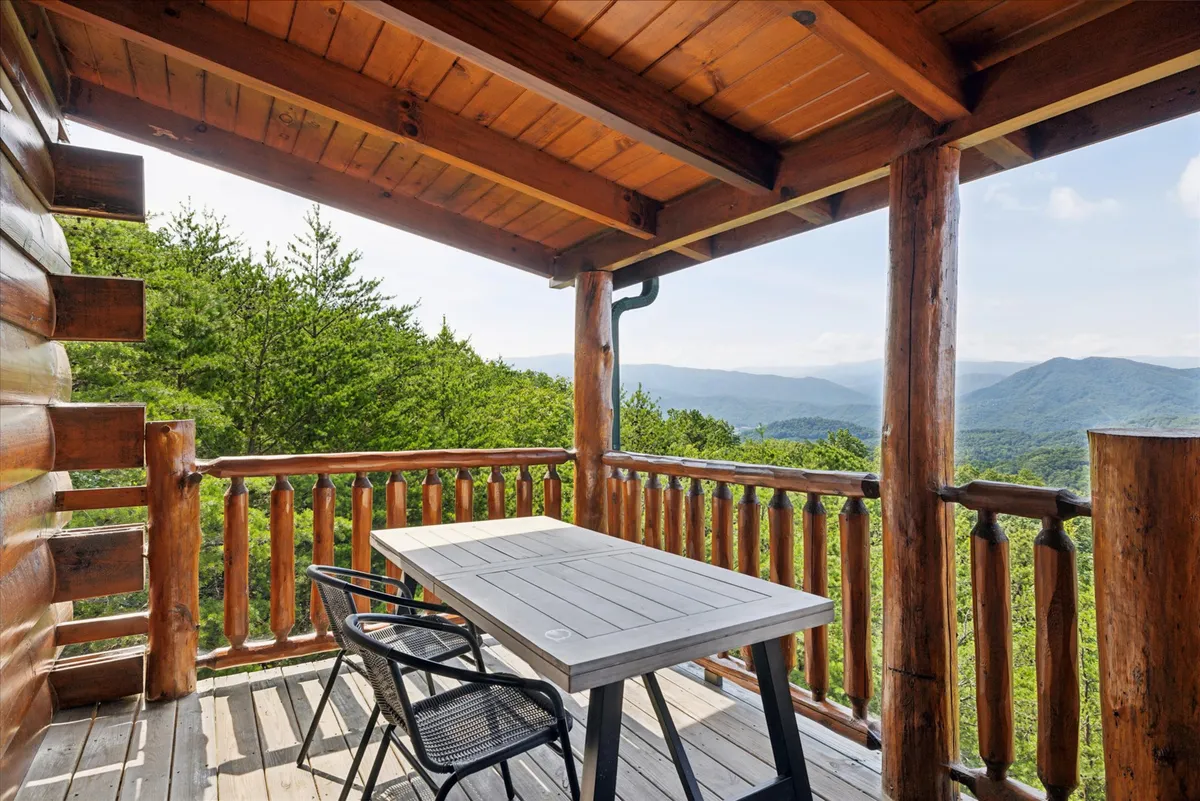 An outdoor table on a wooden deck framed by a sturdy railing faces a panorama of green hills and mountains. The setup invites al fresco dining, laptop work, or quiet reflection, all with the tranquil backdrop of the great outdoors.