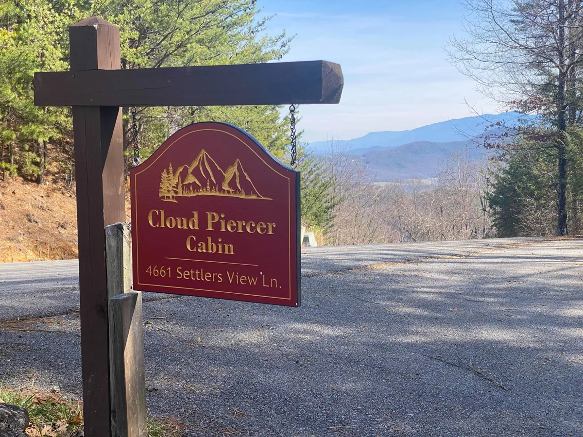 The exterior image features the 'Cloud Piercer Cabin' sign set against a backdrop of rolling mountains and a lush tree line. The visual cues welcome guests to the property while situating it within the broader mountain landscape, hinting at the proximity to local attractions and outdoor adventures.