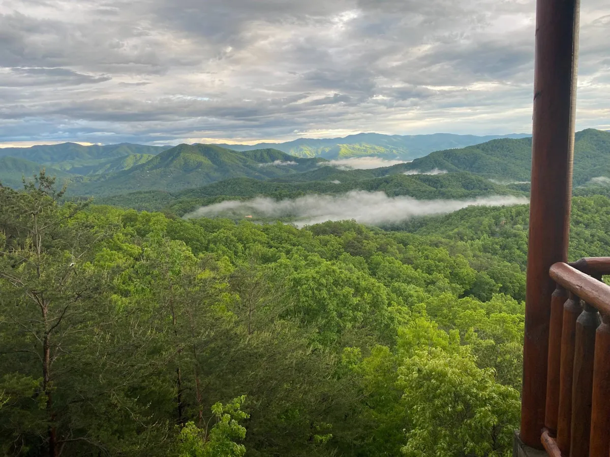 A sweeping view from a balcony or deck, looking out over a vast mountain range, lush green forests, and a dramatic sky. The wooden railing in the foreground ties the property to the untamed wilderness beyond. This immersive perspective invites viewers to picture themselves savoring the scenery after a day of hiking or outdoor excitement.