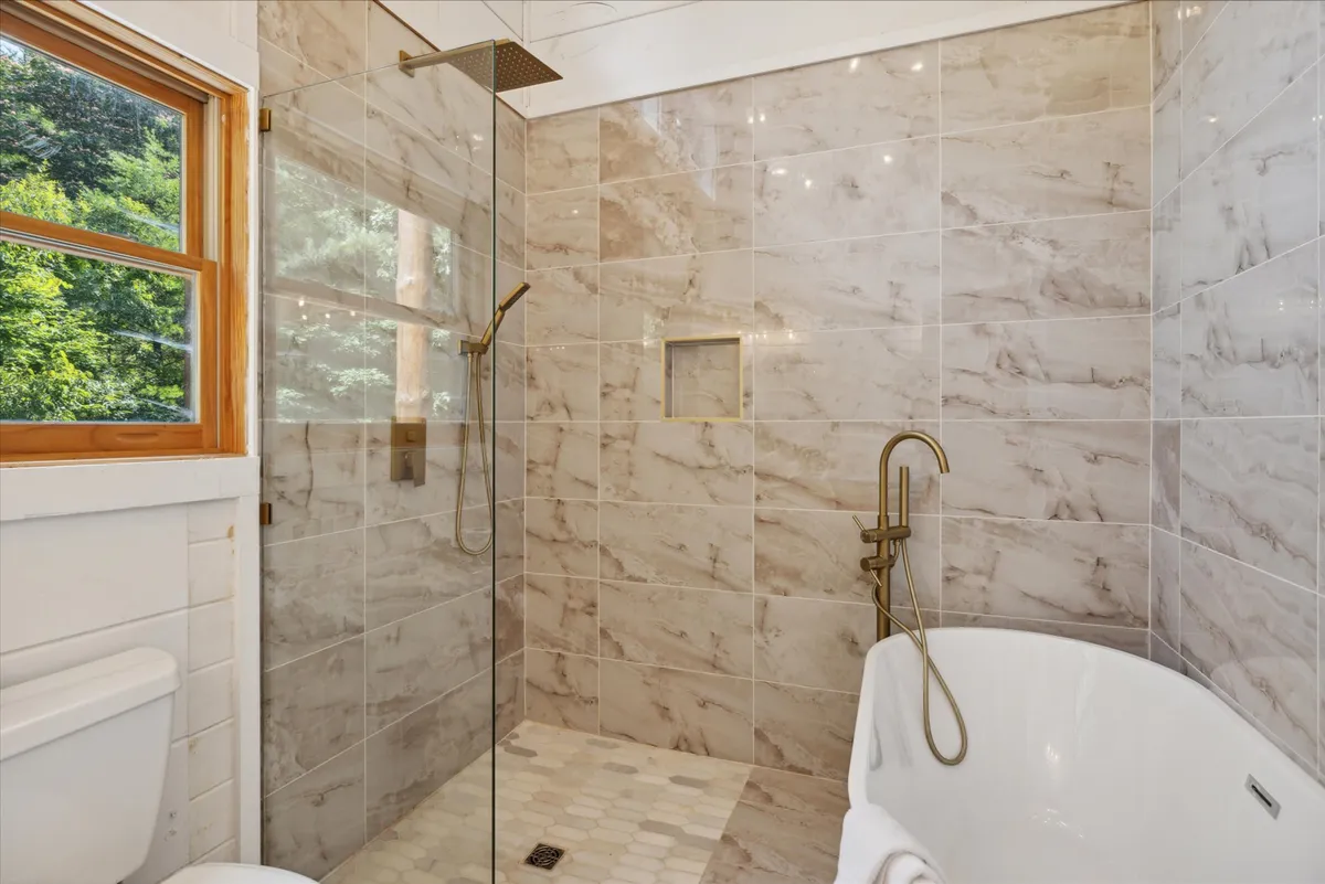 A tranquil bathroom featuring a freestanding bathtub positioned beside a bright window, a glass shower enclosure, and elegant marble tile walls. The space invites relaxation and luxurious self-care.