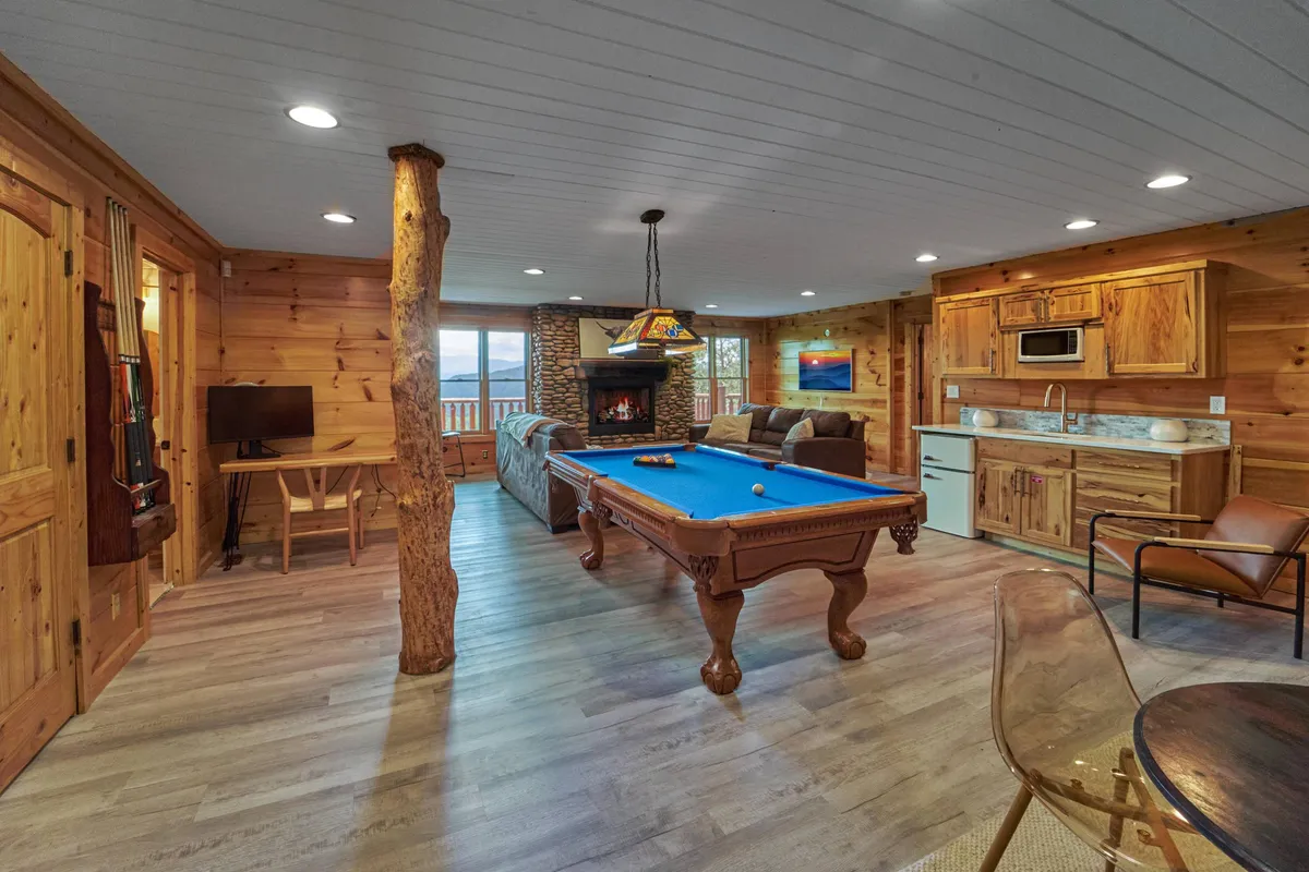 This cozy and inviting interior shows a pool table set before a glowing fireplace, with a kitchen area and large windows that let in natural light. The space is designed for socializing and entertainment, perfect for game nights or relaxing after a workout.