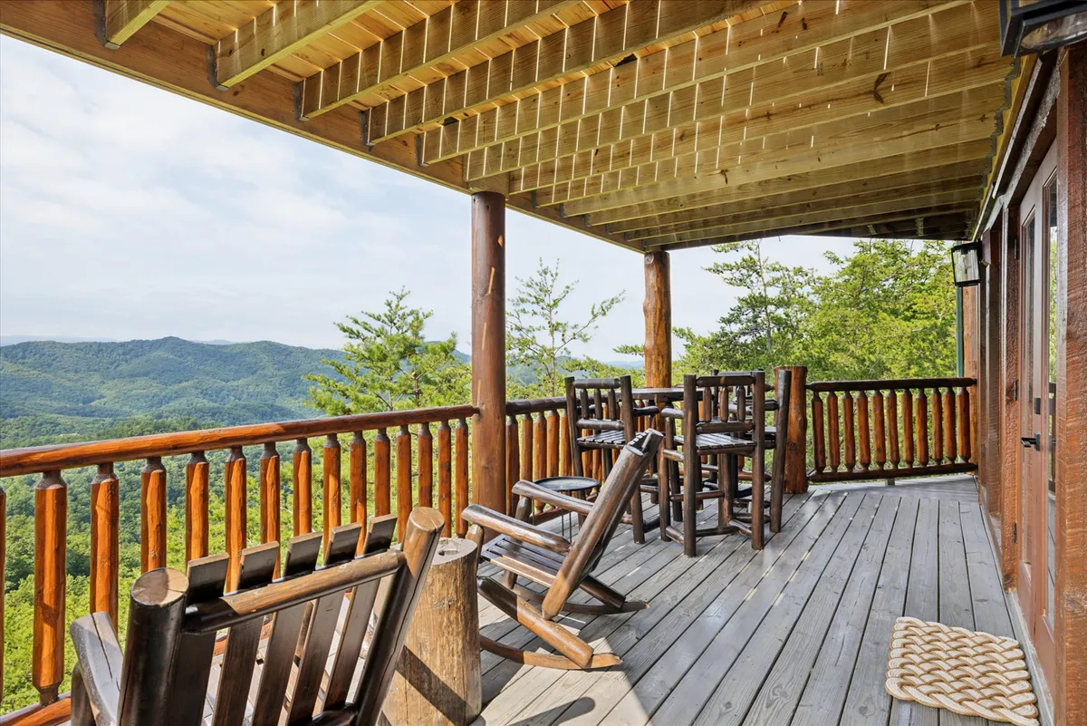 A peaceful exterior view showing a porch with wooden railing, rocking chairs, a table set, and a sweeping mountain view. The welcoming porch invites moments of group relaxation, casual chats, and sunrise or sunset watching for visitors of all ages—including pets at your feet.
