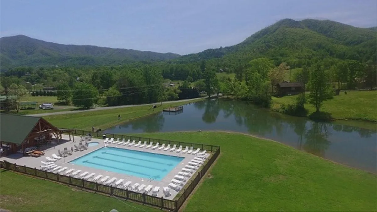 The image showcases the exterior of a luxurious cabin perched above the mountains, with a sparkling swimming pool surrounded by lounge chairs. The breathtaking mountain vista stretches out in the background, and a tranquil lake lies below, all bathed in the golden glow of sunset. The scene radiates relaxation, inviting moments of swimming, sunbathing, and gathering with family or friends against an unforgettable natural backdrop.