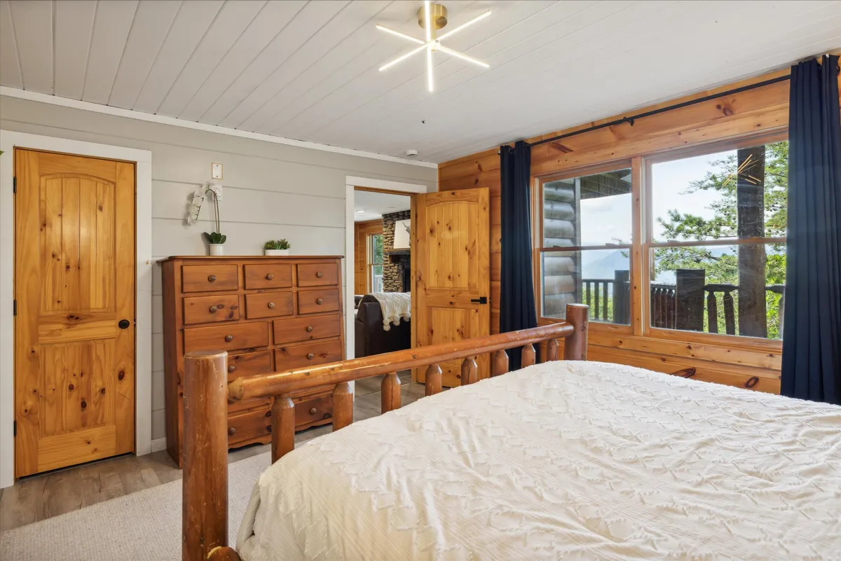 A serene bedroom with a log bed, large windows framed by curtains, and a wooden dresser. The expansive windows fill the room with natural light and offer calming views, enhancing the sense of restful retreat.