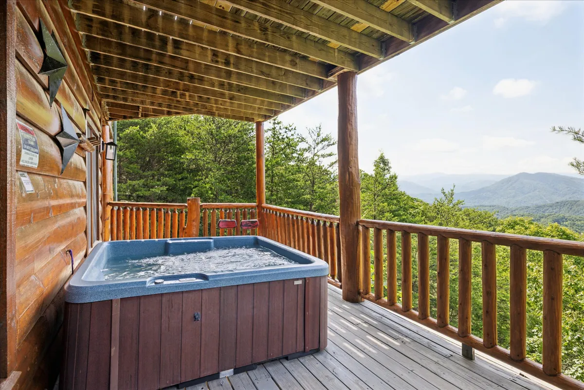 An inviting outdoor hot tub sits on a wooden deck, framed by stunning mountain views and a sturdy railing. Steam rises from the tub, suggesting warmth and relaxation against the cool mountain air.