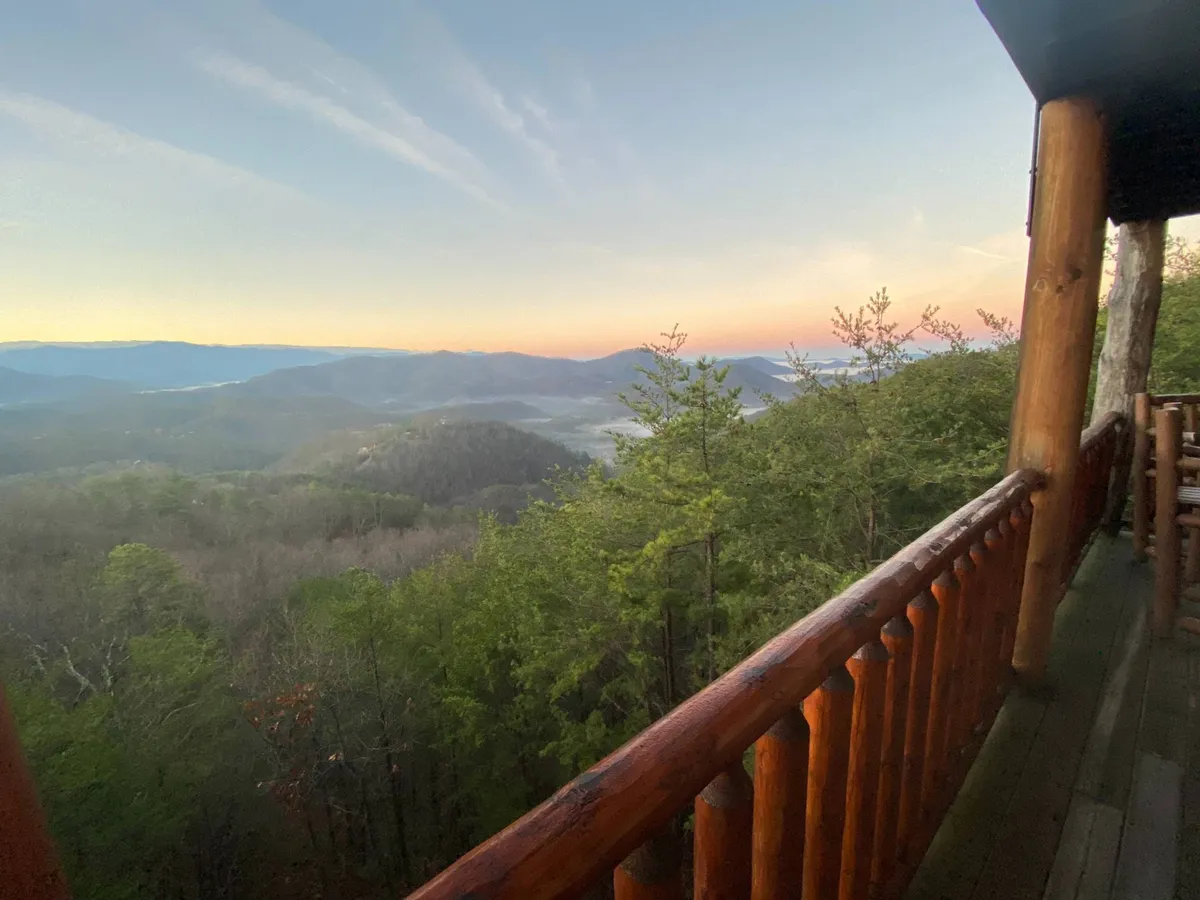 A panoramic view from a wooden porch, featuring a wide stretch of mountains under a colorful sunset sky. The scene invites guests to relax outdoors, enjoy a morning coffee, or simply watch the changing light over the landscape.