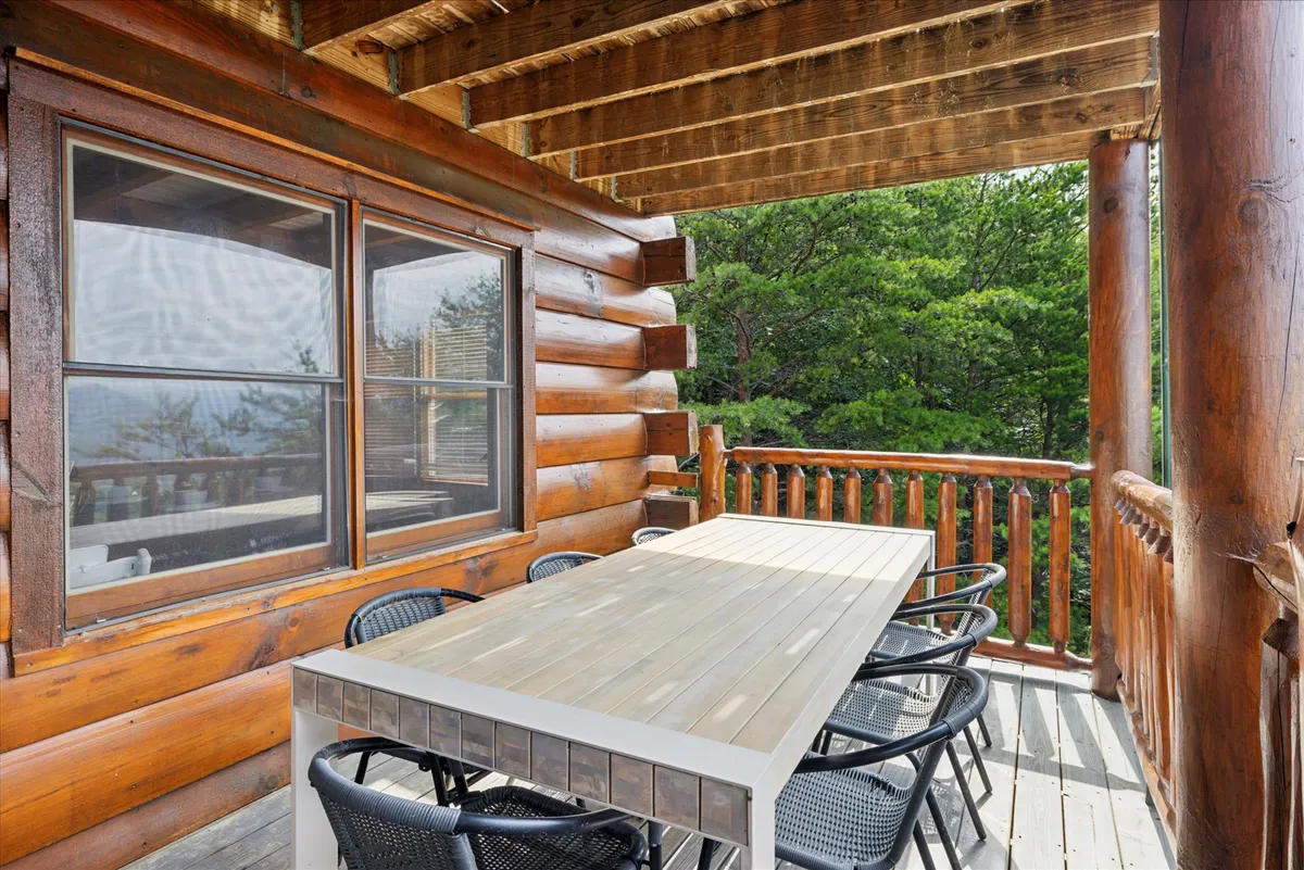 This exterior shot captures a long wooden dining table surrounded by black chairs set on a patio beside the log cabin, with lush greenery all around. The communal table setting is perfect for group gatherings, meals, or relaxed moments with both family and pets, while the open space hints at room for kids and pets to play nearby.