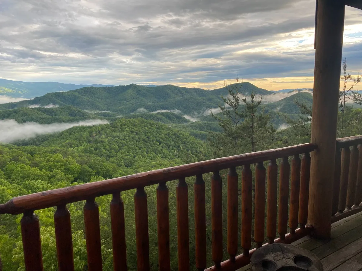 A tranquil mountain landscape stretches out beyond a wooden railing, with clouds drifting overhead and mist hanging in the valleys. The perspective from the deck highlights a peaceful spot perfect for quiet reflection, morning coffee, or stargazing at night.