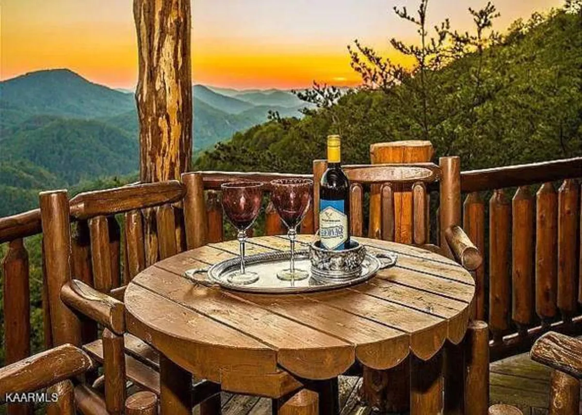 A wooden table set with wine glasses and a bottle sits on a balcony overlooking a sweeping mountain view at sunset. The warm glow of evening light enhances the intimacy of the scene, inviting relaxation and an appreciation of the natural world beyond the railing.
